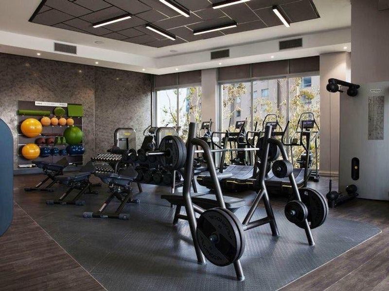 A modern, well-equipped fitness center featuring professional-grade gym equipment, rubberized flooring, recessed linear lighting, and large windows providing ample natural light.