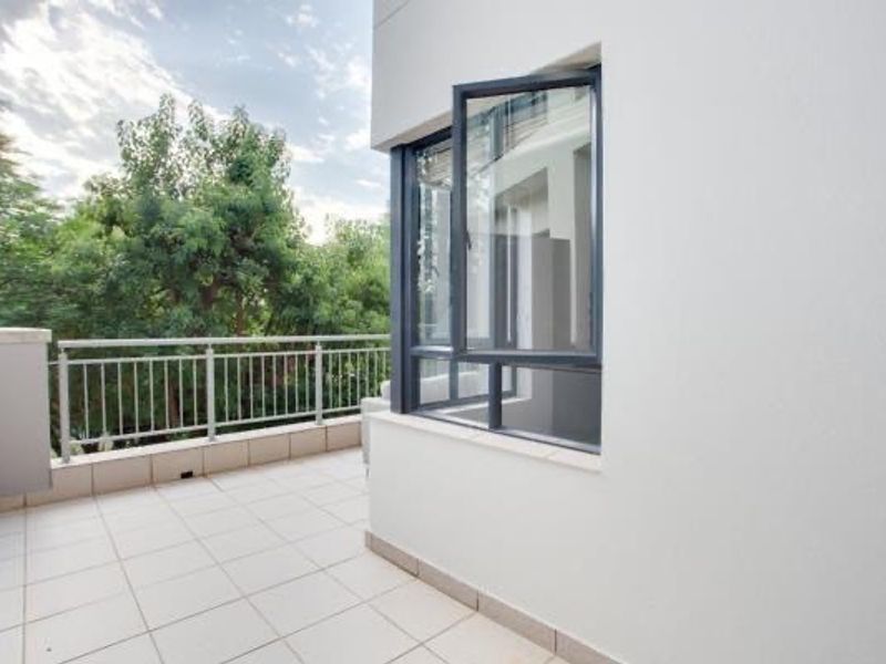 A clean, modern balcony featuring tiled flooring, a secure metal balustrade, and large aluminium-framed windows overlooking lush greenery.