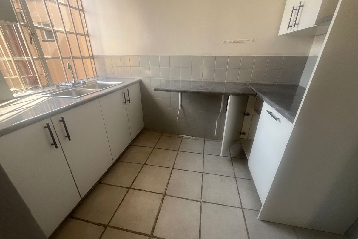 A compact kitchen featuring white cabinetry, laminate countertops, a stainless steel sink, and tiled flooring, currently vacant and in need of minor repairs.
