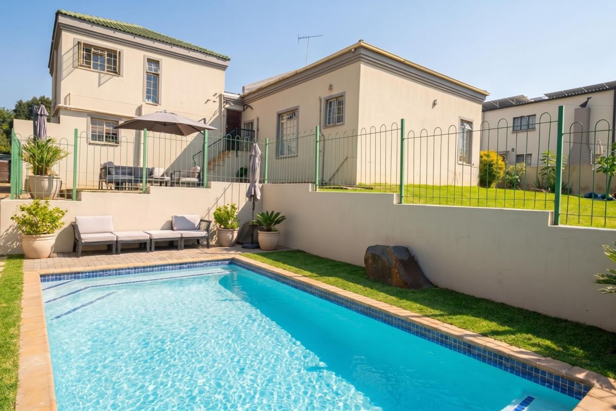 A well-maintained outdoor pool area featuring a clear swimming pool, paved surround, and tiered garden levels enclosed by metal perimeter fencing.