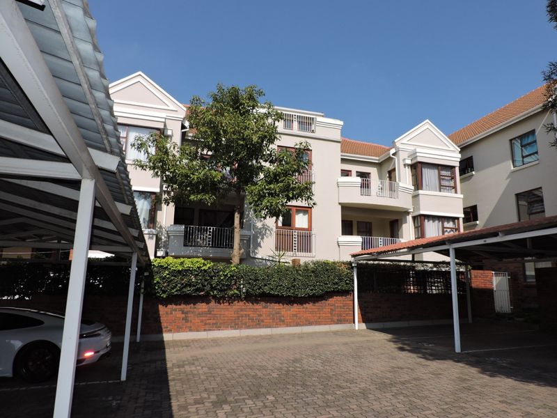 Exterior view of an apartment complex featuring covered parking bays, paved driveway, and multi-story residential units with balconies.