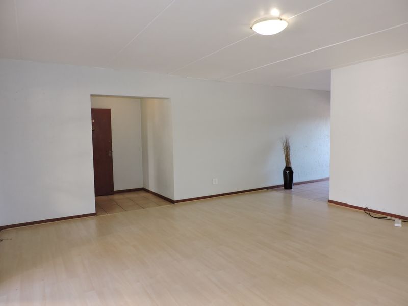 A spacious, empty living area featuring light-toned laminate flooring, neutral walls, and a simple flush-mount ceiling light fixture.