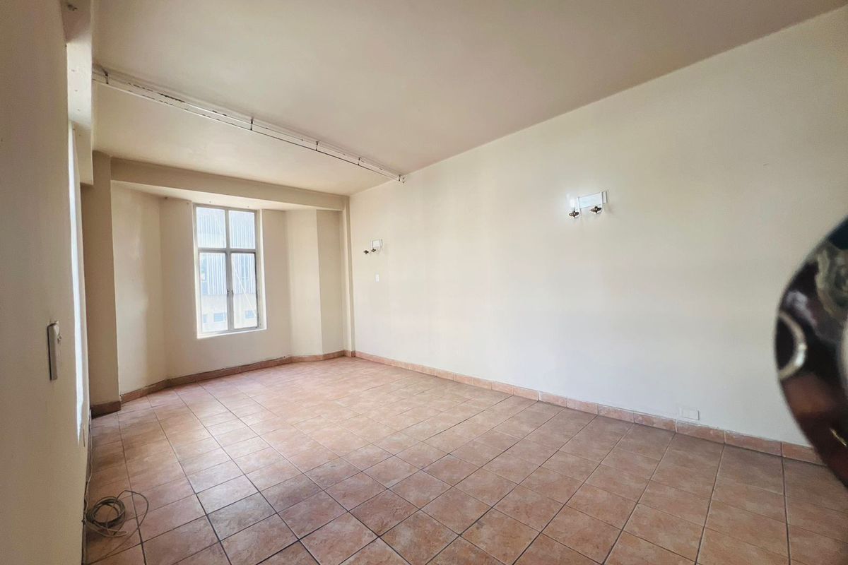 A spacious, empty living area featuring tiled flooring, wall-mounted light fixtures, and a large window providing natural light.