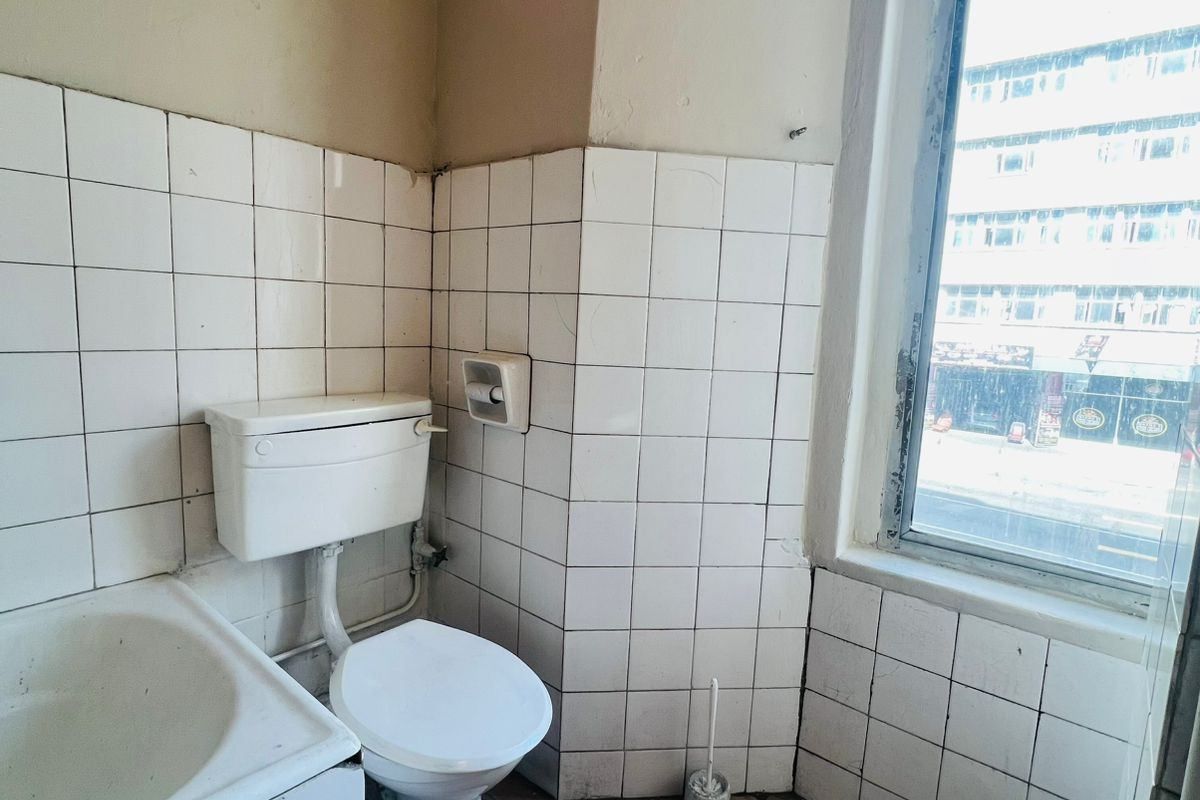 A dated bathroom featuring a tiled bathtub, toilet, and window with street views. The space requires modernization of finishes and fixtures.