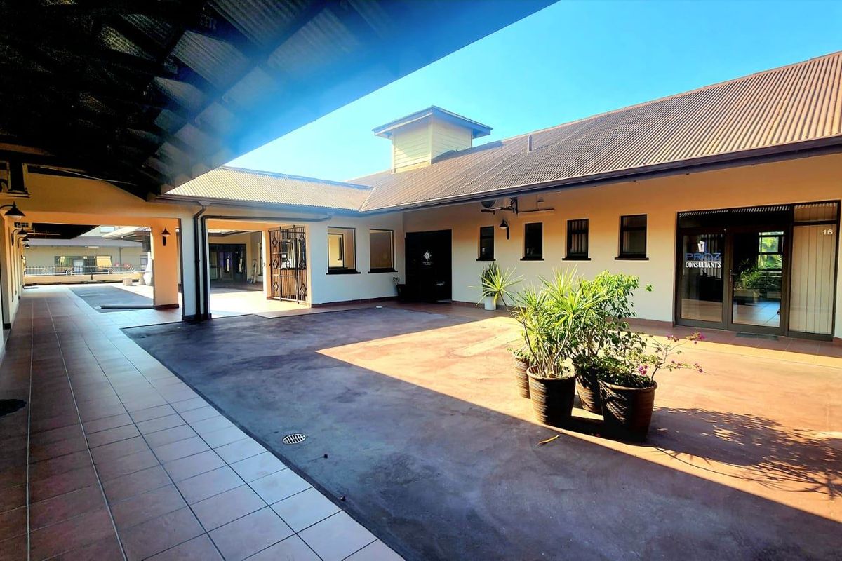 A spacious, paved commercial courtyard featuring a covered walkway, tiled flooring, and multiple office access points under a metal roof structure.