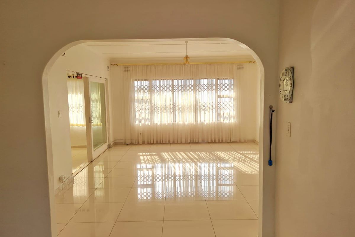A bright, empty living room featuring polished tiled flooring, an arched entryway, and a large window with security gate and sheer curtains.