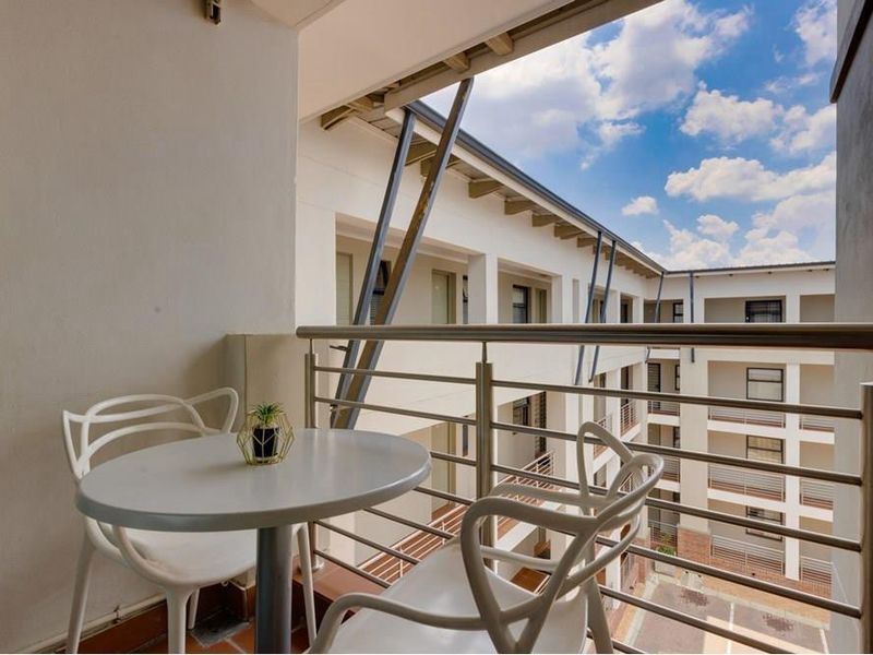 A covered balcony featuring a stainless steel balustrade and tiled flooring, overlooking the apartment complex courtyard.