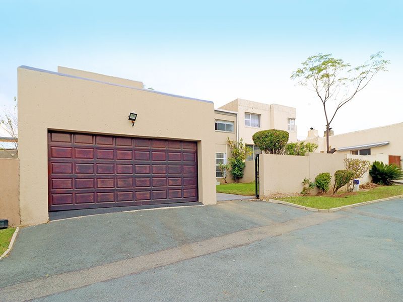 Front exterior view of a cluster home featuring a double garage, paved driveway, and boundary wall with established garden elements.