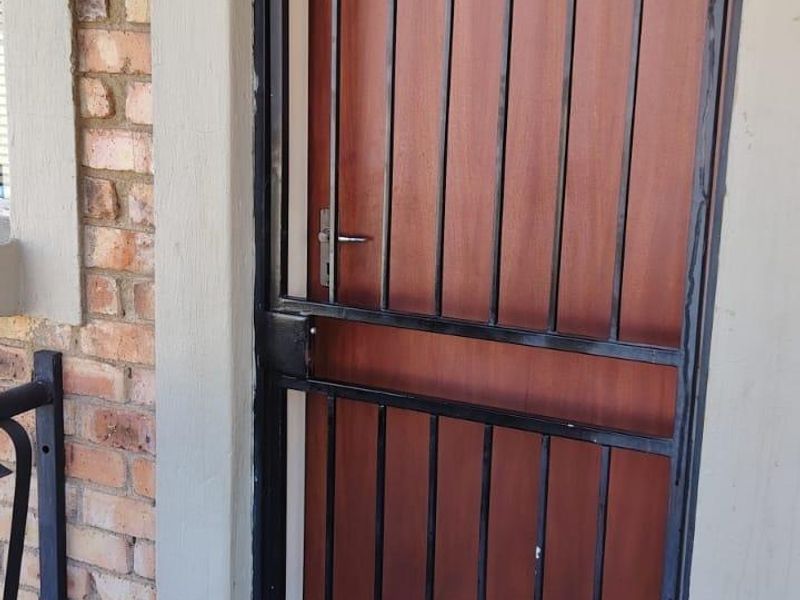 Front entrance featuring a wooden door with a black metal security gate, set against a brick facade with tiled porch flooring.