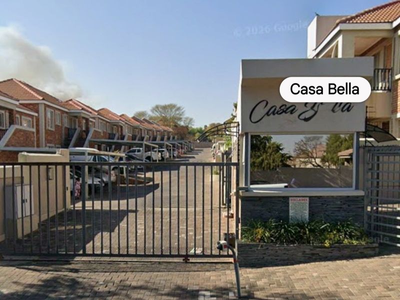 Entrance view of Casa Bella townhouse complex featuring a secure gated access point, paved driveway, and rows of residential units with carports.