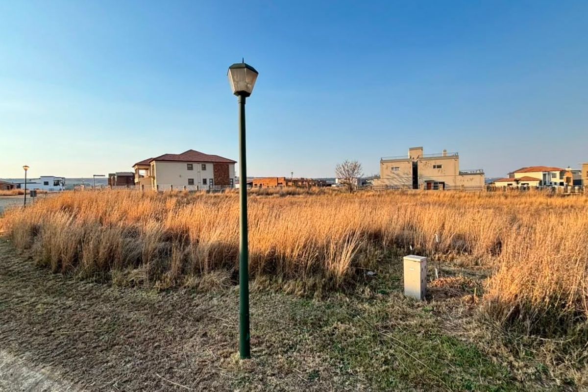 A vacant residential plot featuring tall dry grass, a street lamp, and utility connection point, situated within a developed residential estate.