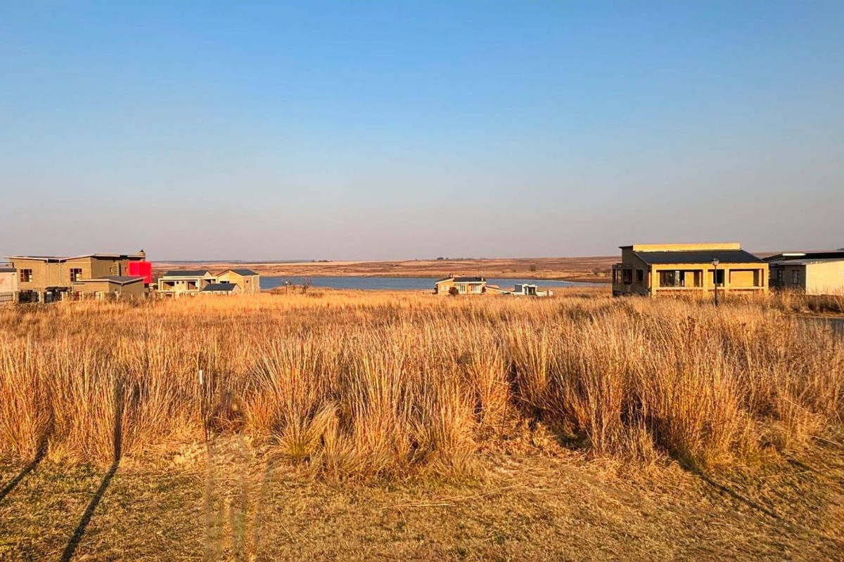 A wide-angle view of a vacant residential plot featuring natural dry grass vegetation with surrounding estate homes and a distant water view.