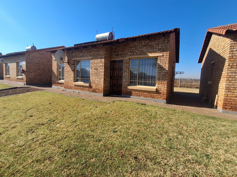 A well-maintained face-brick townhouse exterior featuring a tiled roof, solar geyser, security gate, and a neat front lawn with a paved walkway.