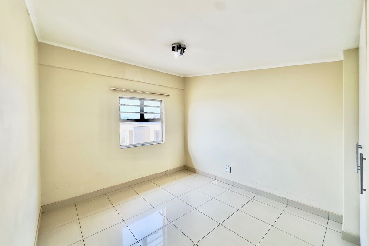 A bright, empty bedroom featuring neutral tiled flooring, a built-in cupboard, and an aluminium-framed window.