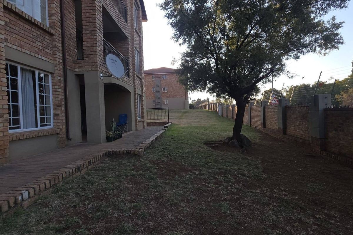 Rear view of a face brick apartment building featuring a paved patio area, balcony, and a spacious garden with perimeter wall and electric fencing.