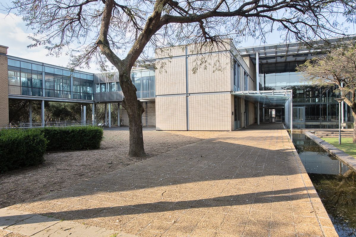 A modern commercial building exterior featuring glass curtain walls, brick cladding, a covered walkway, and a landscaped courtyard with a water feature.
