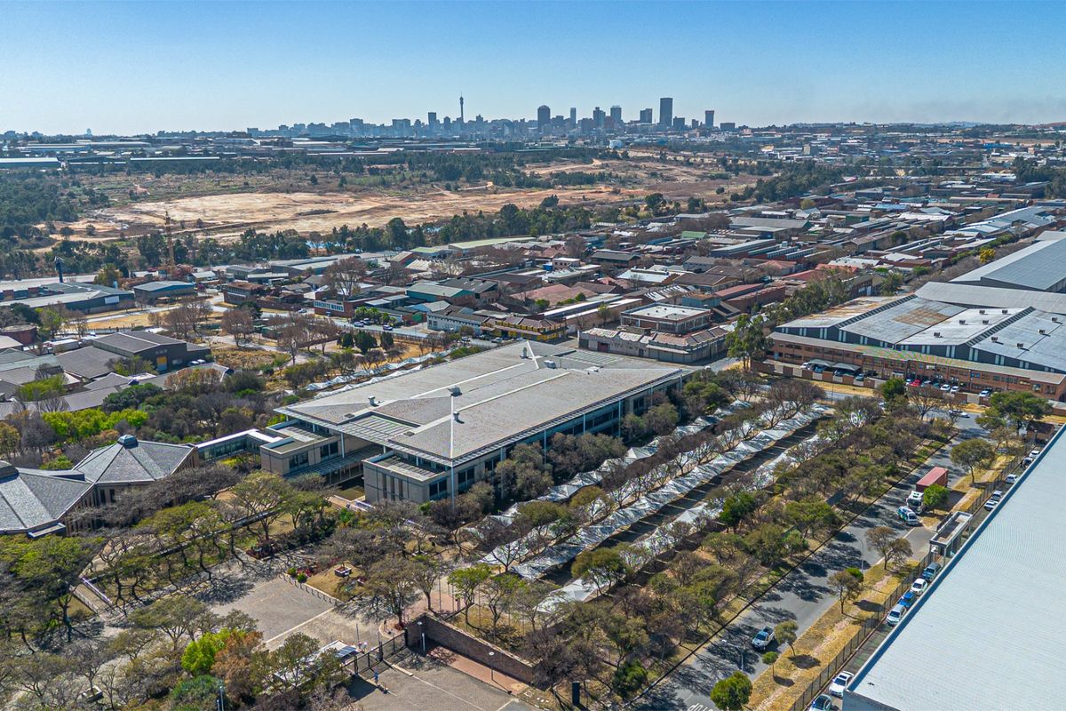 Aerial view of a commercial property complex featuring extensive parking facilities, landscaped grounds, and surrounding industrial infrastructure with a city skyline in the distance.