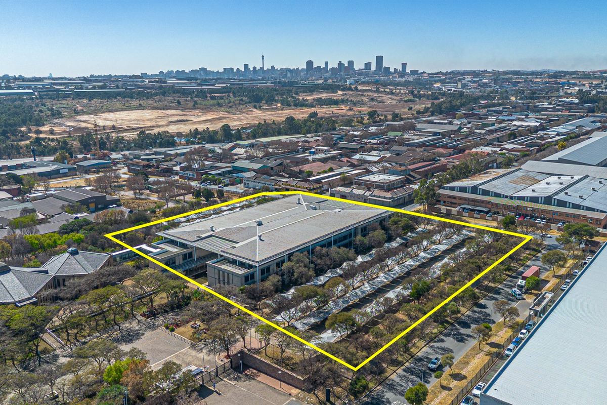 Aerial view of a commercial property featuring a large office building, extensive parking facilities, and landscaped grounds with city skyline views in the distance.