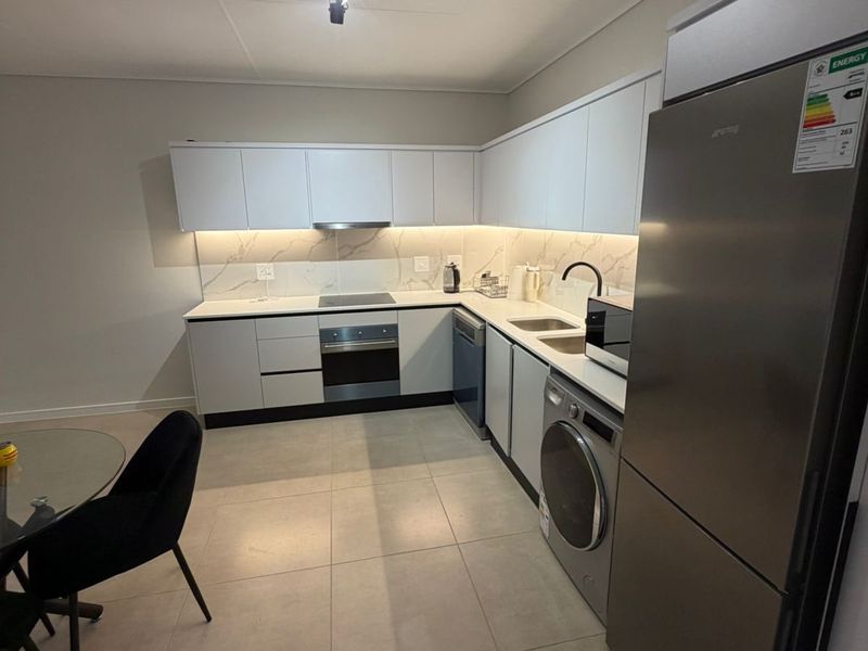 Modern kitchen featuring sleek white cabinetry, stone-look backsplash, integrated oven, electric hob, and stainless steel appliances with neutral tiled flooring.