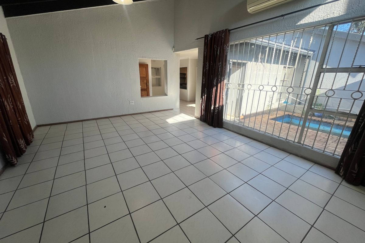 Spacious, light-filled living area featuring tiled flooring, exposed ceiling beams, and a large window with security bars overlooking the pool area.