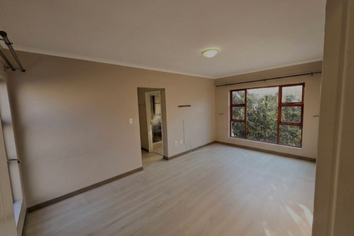 A bright, empty bedroom featuring light-toned laminate flooring, neutral walls, and large timber-framed windows overlooking greenery.