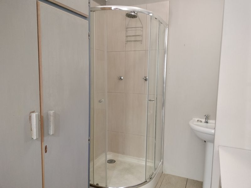 A functional bathroom featuring a curved glass shower enclosure, pedestal basin, and built-in storage cupboard with neutral wall tiling.