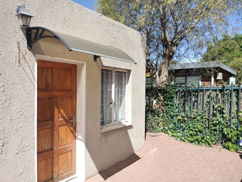 Rear exterior view of the apartment featuring a textured plaster facade, wooden entrance door with canopy, security window bars, and a paved patio area.
