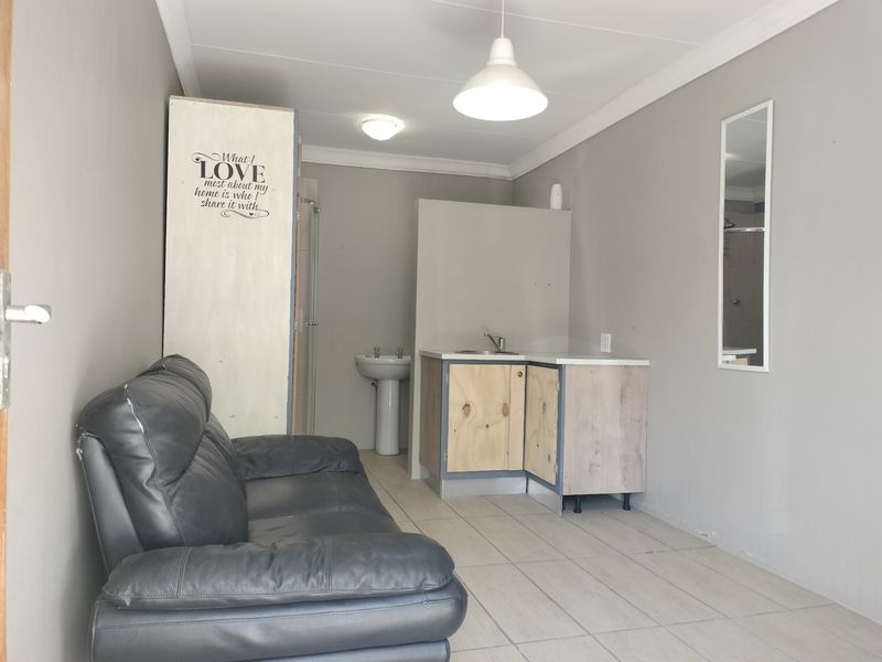 Open-plan living area featuring a basic kitchenette with sink and a separate pedestal basin, finished with neutral wall tones and tiled flooring.