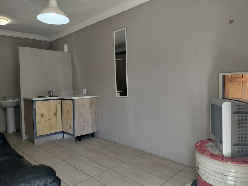 Open plan living area featuring a basic kitchenette with sink and a separate pedestal basin, finished with tiled flooring and neutral wall paint.