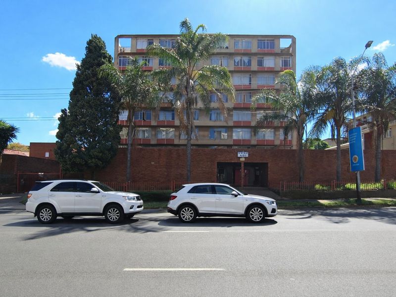 Street view of a multi-story apartment building featuring a brick perimeter wall and mature palm trees.