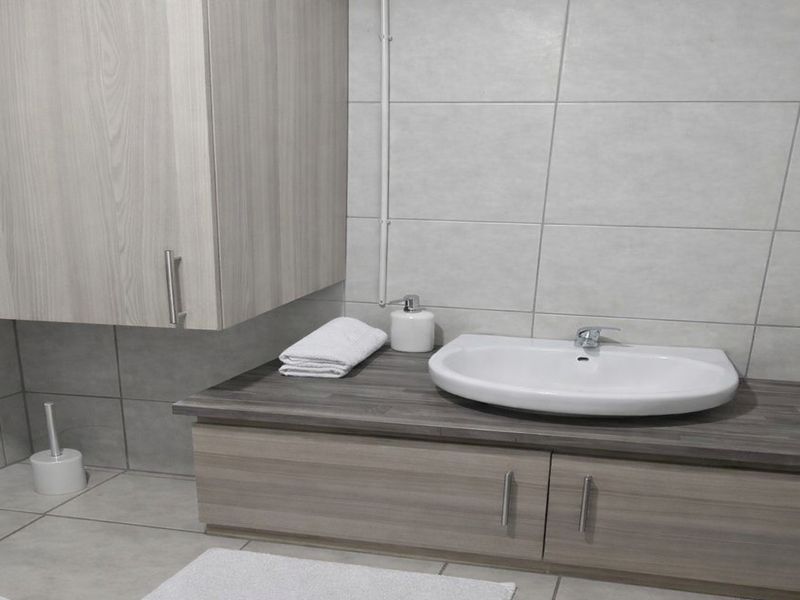 A modern bathroom vanity featuring a wood-grain cabinet, dark laminate countertop, and a white ceramic basin with wall-mounted tiling.