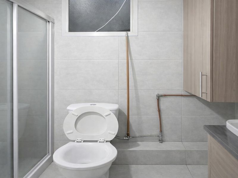 A functional bathroom featuring a shower enclosure, toilet, and vanity cabinet with exposed copper plumbing fixtures.