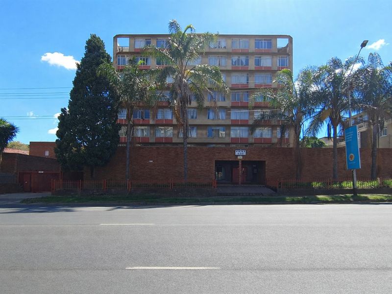 Street view of a multi-story apartment building featuring a brick perimeter wall, gated entrance, and established palm trees.