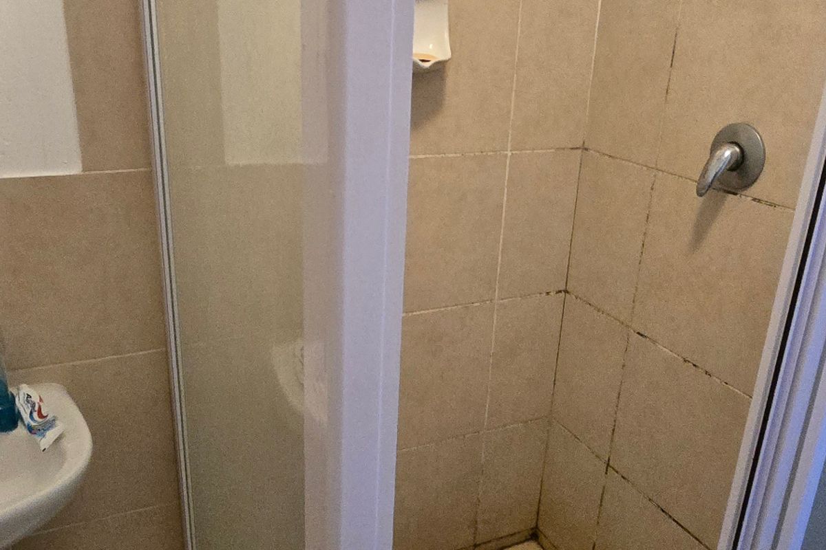 A compact bathroom shower enclosure featuring tiled walls and flooring, requiring maintenance to address grout discoloration and surface grime.