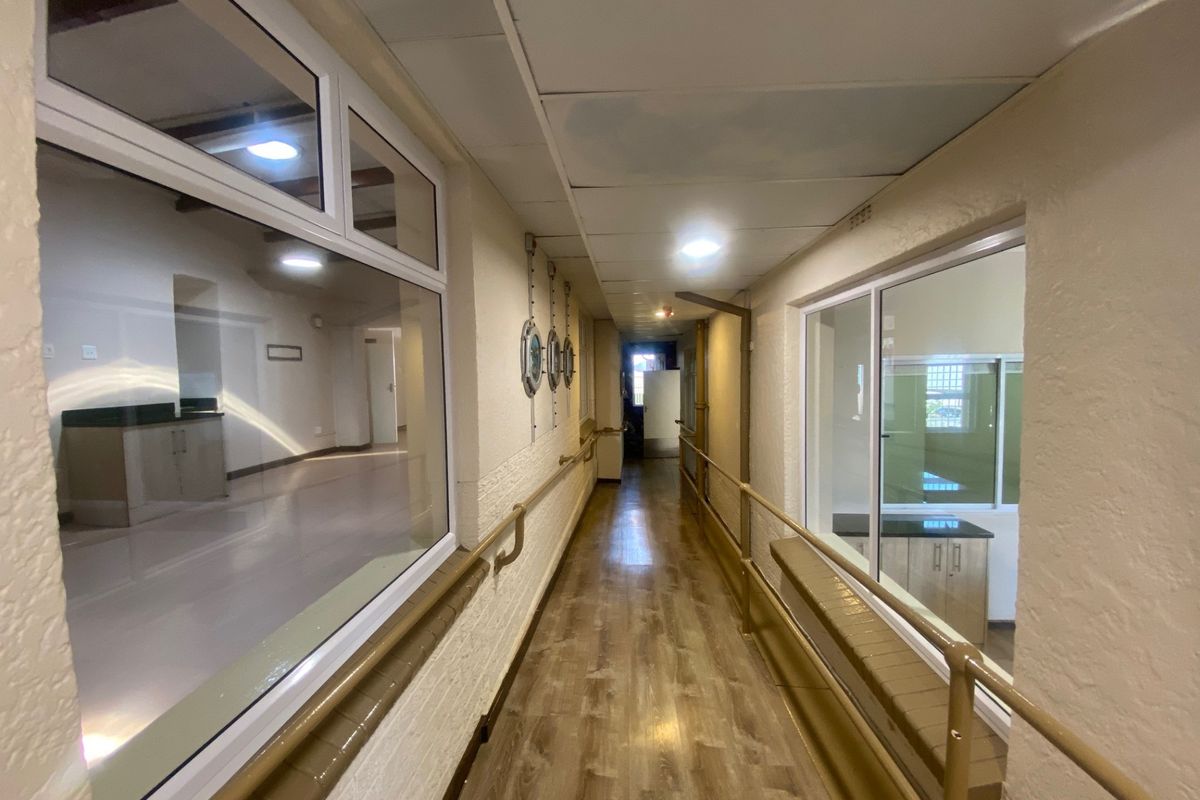 A long, well-lit commercial hallway featuring laminate flooring, handrails, and large glass partitions looking into adjacent office or workspace areas.