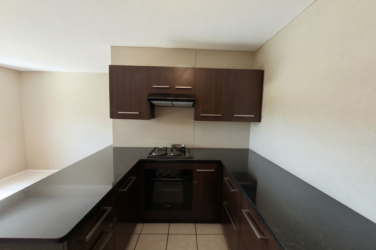 A compact, modern kitchen featuring dark wood cabinetry, granite countertops, a built-in oven, gas hob, and extractor fan with neutral tiled flooring.