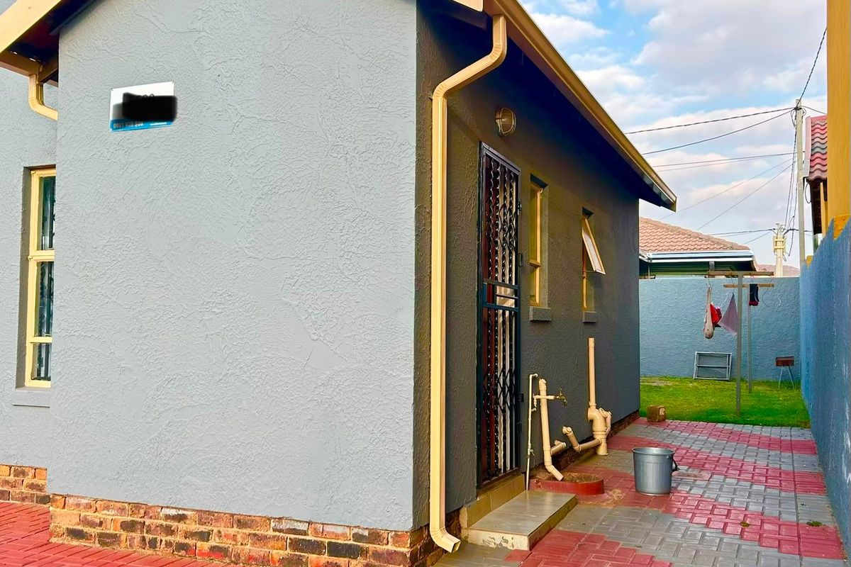 A view of the rear exterior of the house featuring paved ground, security gate, and external plumbing fixtures.