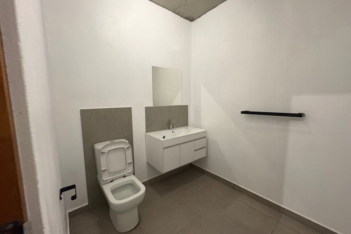 A modern, minimalist bathroom featuring a wall-mounted vanity, ceramic toilet, tiled flooring, and an exposed concrete ceiling, presenting a clean and contemporary aesthetic.