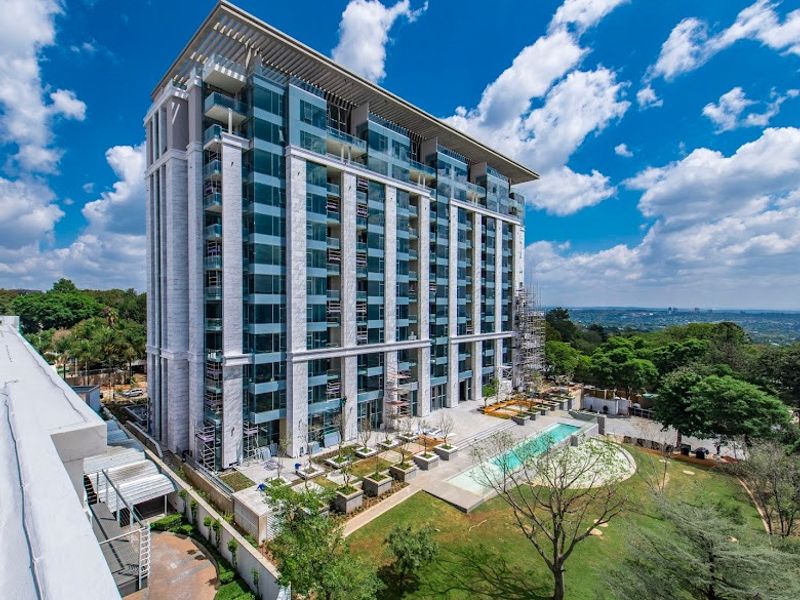 Aerial view of a modern high-rise apartment building featuring glass balconies, a swimming pool, and landscaped garden areas.