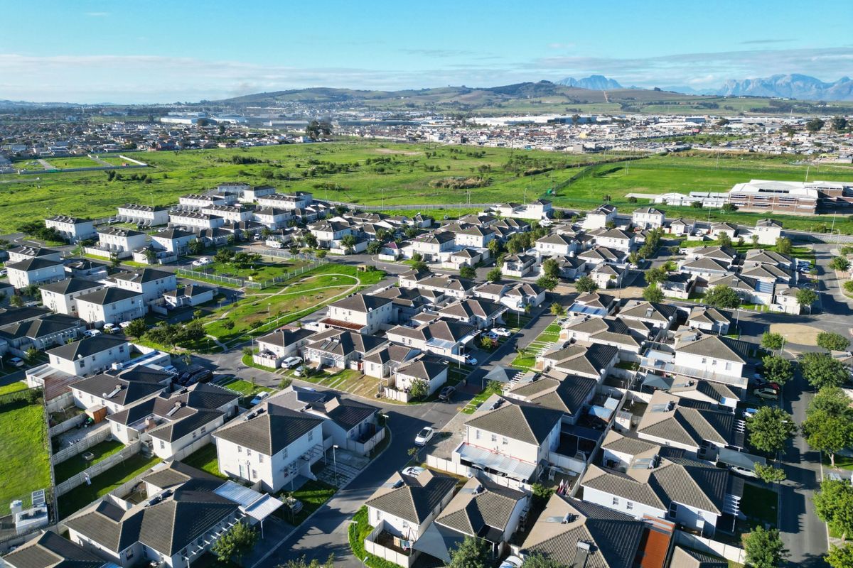 An aerial view of a modern residential estate featuring uniform housing units, paved streets, and surrounding green space with distant mountain views.