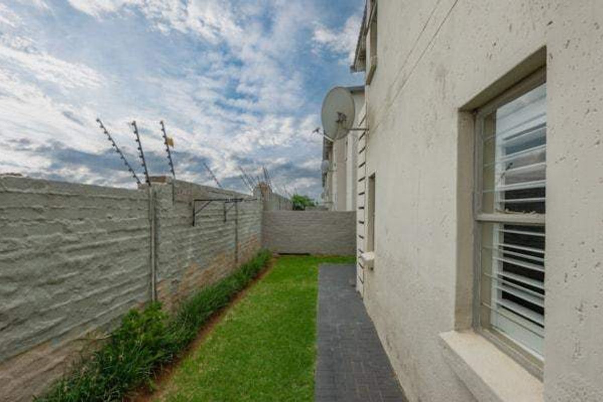 A side garden area featuring a paved walkway, lawn, boundary wall with electric fencing, and security bars on the windows.