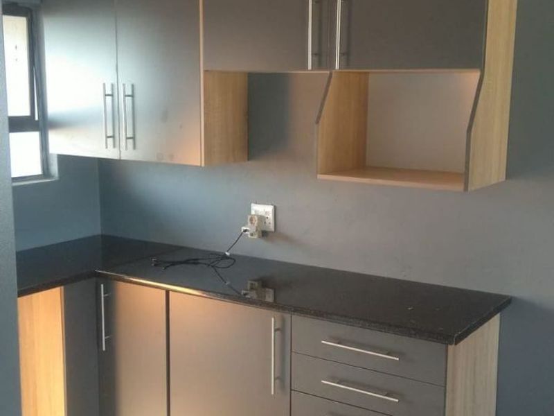 A modern, compact kitchen featuring grey and wood-tone cabinetry, dark granite countertops, and neutral tiled flooring.