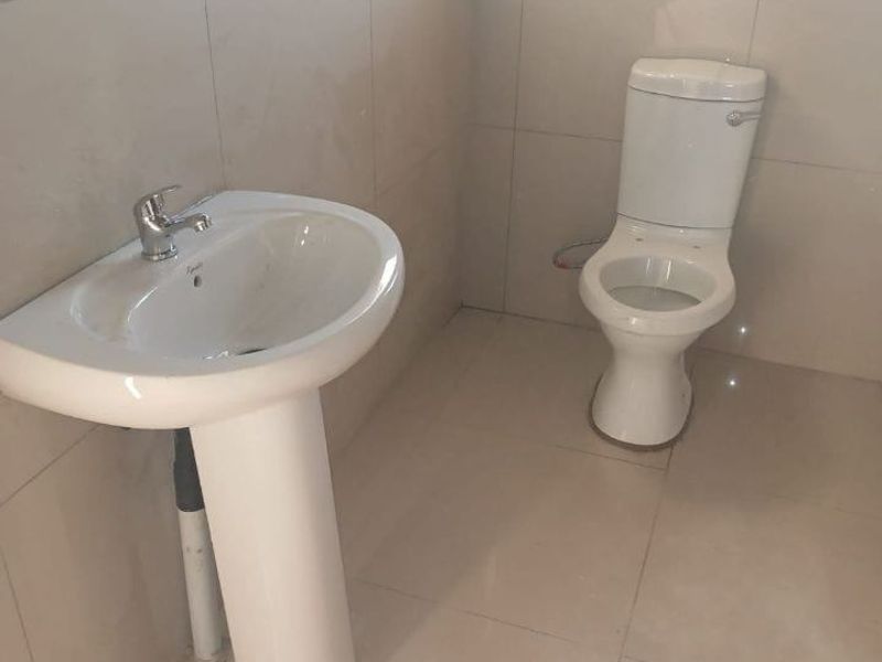 A clean, minimalist bathroom featuring a white pedestal basin and a standard toilet, finished with neutral ceramic wall and floor tiles.