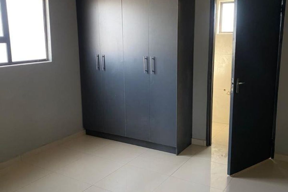 A clean, empty bedroom featuring modern dark built-in cupboards, neutral tiled flooring, and a panelled ceiling, with an adjacent en-suite bathroom.