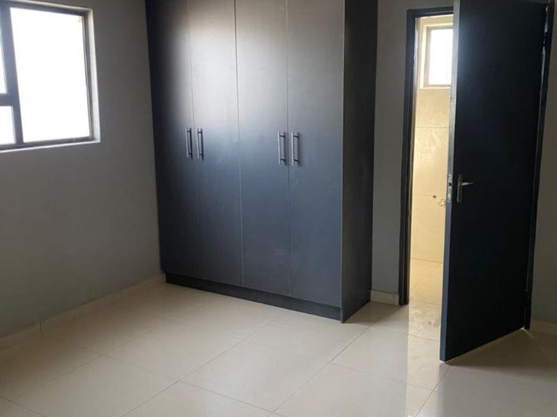 A clean, empty bedroom featuring modern dark built-in cupboards, neutral tiled flooring, and a panelled ceiling, with an adjacent en-suite bathroom.