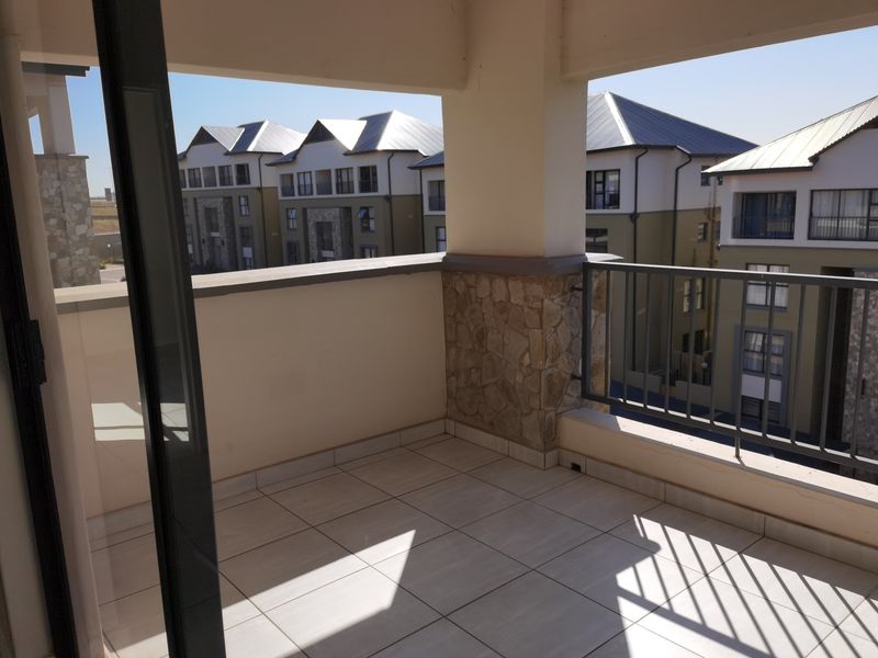 A covered balcony featuring tiled flooring, stone-clad pillars, and a metal balustrade, offering an open view of the surrounding apartment complex.