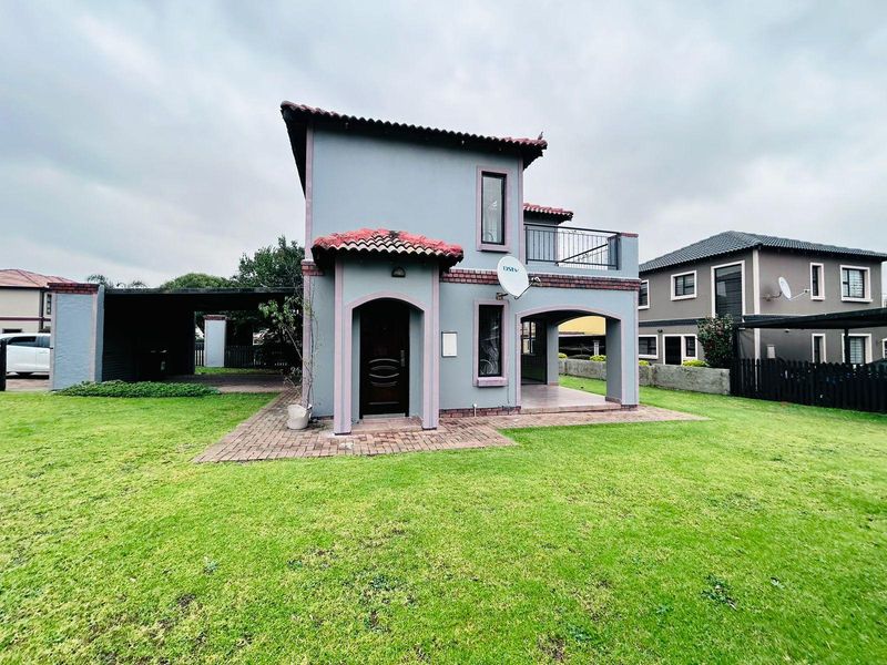 Rear view of a two-story townhouse featuring a covered patio, upper balcony, tiled roof, and an attached carport set within a spacious garden.