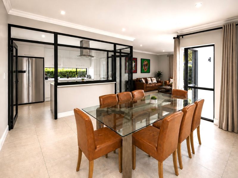 Modern open-plan living and dining area featuring a sleek kitchen enclosed by a stylish black-framed glass partition, leading to a bright, tiled living space.