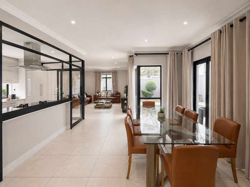 Spacious open-plan living and dining area featuring a modern black steel-framed glass kitchen partition, tiled flooring, and seamless access to the outdoor patio.