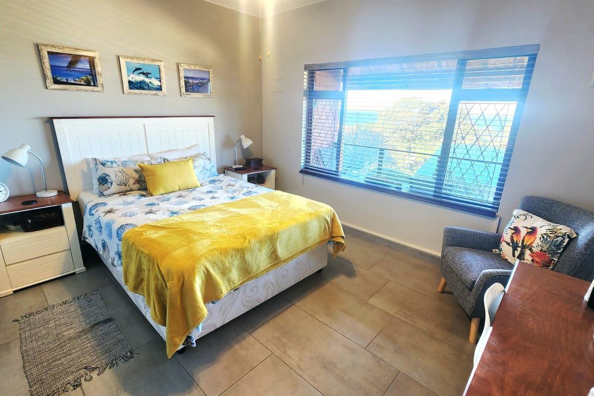 A well-lit bedroom featuring neutral wall tones, tiled flooring, and a large window with venetian blinds offering natural light and exterior views.